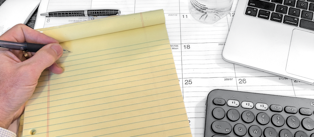 Photo of a somewhat messy desk, with keyboard and calendar, and a yellow legal pad in the foreground. A hand holding a pen is poised to write on the legal pad